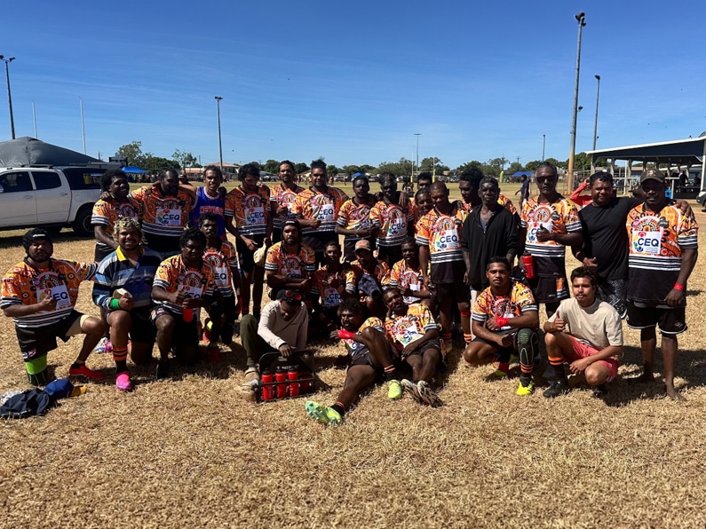Group of rugby players in orange and black jerseys posing on a sunny field front row seated with water bottles and gear visible